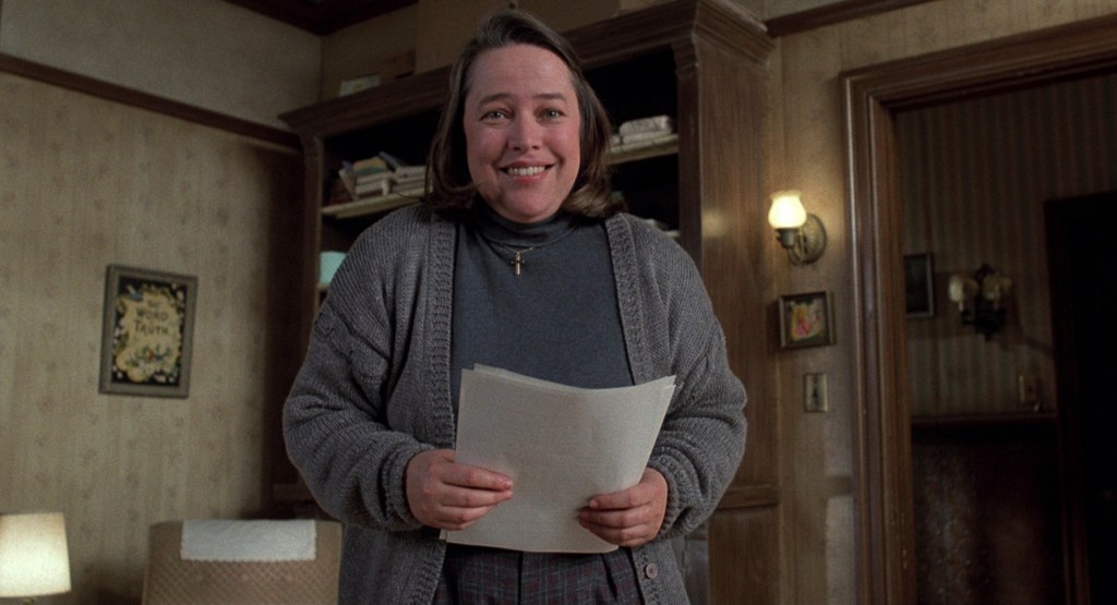 Kathy Bates in Misery. She stand center frame, a smile on her face, and papers in her hands. 