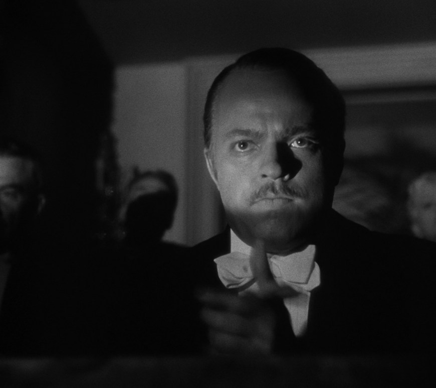 Still from Citizen Kane. Orson Welles as Kane sits in darkness, light showing his eyes. He claps.
